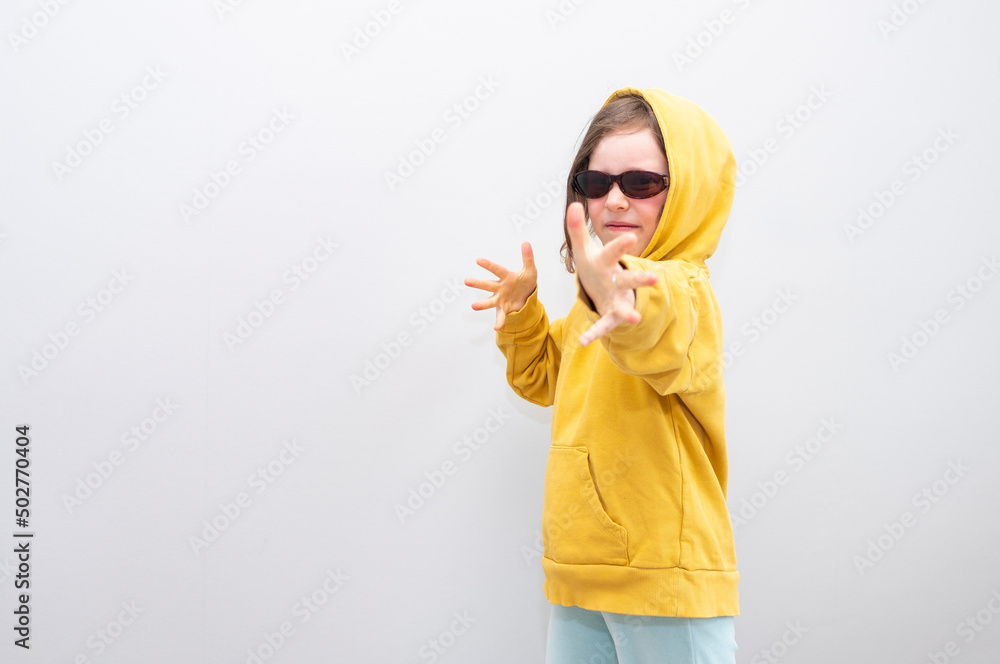 Caucasian girl 6 years old, in a yellow sweatshirt with a hood on a gray background. The child smiles and poses for the camera, showing a shot from a bow with hand gestures. Caucasian girl 6 years old, in a yellow sweatshirt with a hood on a gray background. The child smiles and poses for the camera, showing a shot from a bow with hand gestures.