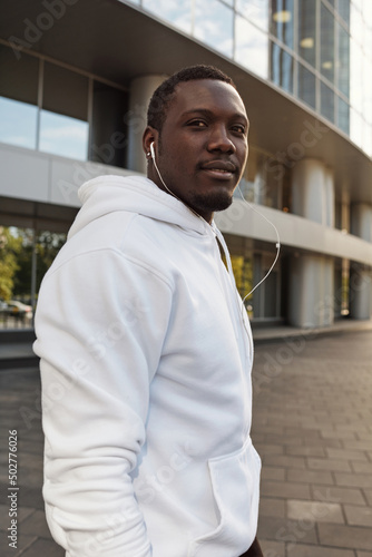 Portrait of young confident Black man in white hoodie and earphones standing outdoors against modern business center and smiling at camera