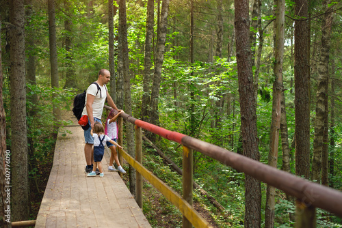 Dad and daughters are standing on old wooden footbridge in woods among trees. Hiking with whole family with small kid. Traveling through ecological trail. Man with hiking backpack holds girls by hands