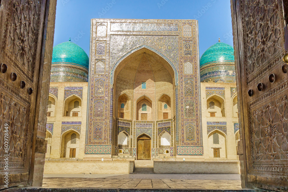 View of the Miri Arab madrasah from the open old wooden doors in Bukhara, Uzbekistan. Perfect sunny day and fairy tale architecture seem to invite you to walk along the ancient streets
