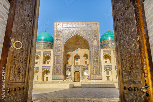View of the Mir-i Arab madrasah from the open ancient wooden doors in Bukhara, Uzbekistan. Sunny day and magnificent architecture seem to invite you to walk along the old streets
