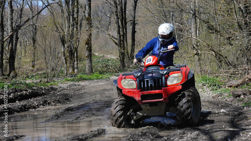 A girl driving a red quad bike rides through a puddle in the spring forest. Front view. Extreme type of outdoor activities. ATV riding.