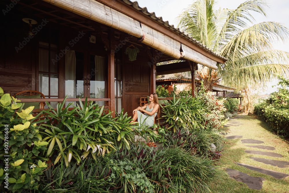 Attractive woman sitting on veranda of wooden house, relaxed summery atmosphere. Vacation on Bali, Ubud. Tropical view on background, palm tree and rice terraces Attractive woman sitting on veranda of wooden house, relaxed summery atmosphere. Vacation on Bali, Ubud. Tropical view on background, palm tree and rice terraces