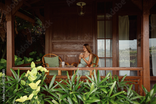 Girl drinking coffee / tea and enjoying the sunrise / sunset on wooden house terrace with garden view