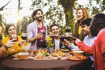 Group of friends dining outdoors toasting with red wine in the backyard - multi generational diverse people having fun together in the nature sitting around the dinner table - focus on the mature man
