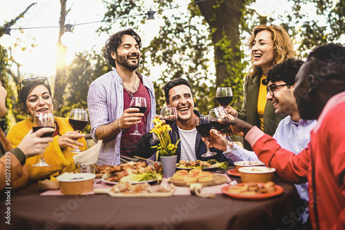 Group of friends dining outdoors toasting with red wine in the backyard - multi generational diverse people having fun together in the nature sitting around the dinner table - focus on the mature man