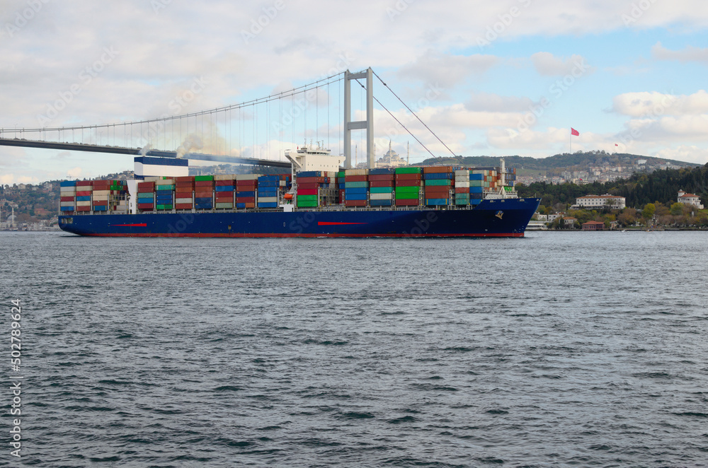 A cargo ship in the Bosphorus. Container ship with rows of containers. Concept of shipping containers by vessel. Cityscape with bridge over Bosphorus in the background