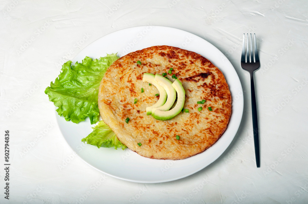 traditional Swiss dish is a potato pancake on a white plate decorated with a leaf of green lettuce and avocado, with a fork next to the plate.
