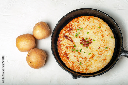 traditional Swiss dish is a potato pancake in a frying pan, decorated with green onions. whole tubers of unpeeled potatoes lie nearby. top view.