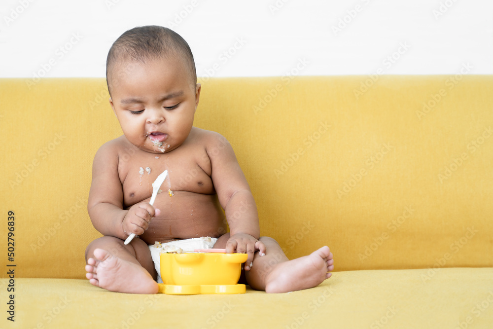 Adorable African baby newborn in diaper sitting on yellow sofa trying to grab a spoon to feed herself. Spilled food messy in mouth and body of little kid. Child development concept Adorable African baby newborn in diaper sitting on yellow sofa trying to grab a spoon to feed herself. Spilled food messy in mouth and body of little kid. Child development concept