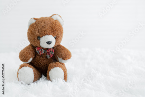 Small brown bear doll with red and green plaid pattern bow placed on fluffy carpet. White background. Copy space