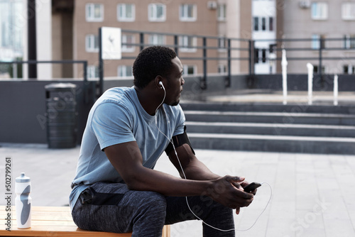 Young athletic African American man in sportswear resting on bench in city center and listening to music through earphones after workout