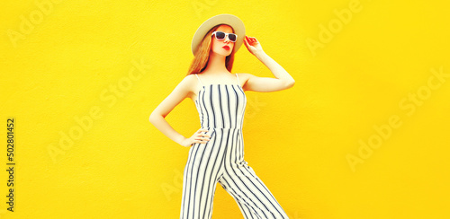 Stylish model woman wearing summer round straw hat, white striped jumpsuit posing on yellow background