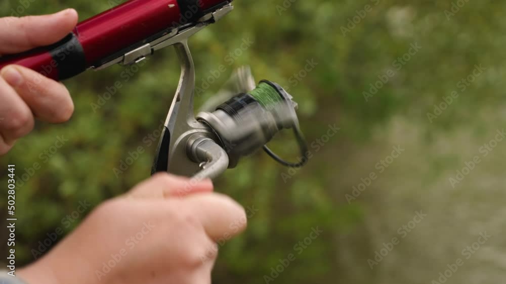 A woman's hand turns the reel handles on a red fishing rod. Close-up. Green leaves blurred in the background. The concept of recreation and hobbies in nature A woman's hand turns the reel handles on a red fishing rod. Close-up. Green leaves blurred in the background. The concept of recreation and hobbies in nature