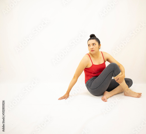 athletic woman in gray sportswear, red blouse, sitting contemplating, with open eyes and holding one leg, on white background