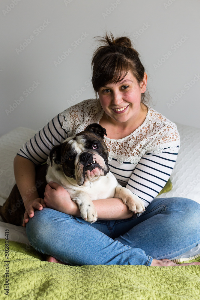 Pretty brunette young woman in casual clothes holding her pet British Bulldog while sitting cross-legged on bed