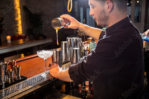Serious young barman preparing cocktail in shaker in bar