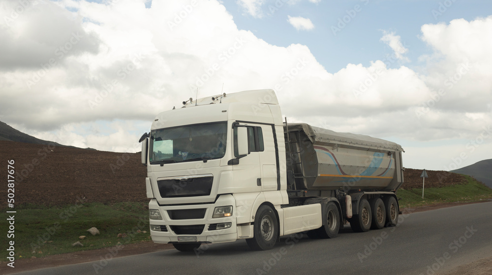 A big truck with a white trailer with space for text on a countryside road in motion against a blue sky with clouds