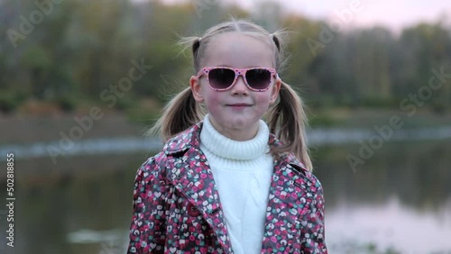 Portrait of a fashionable cute Caucasian child girl who is wearing stylish sunglasses. A little girl stands alone in a city public park, looks at the camera and smiles. Fashion and lifestyle concept