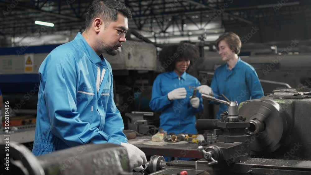 Selective focus of Asian male lathe worker in a blue uniform and gloves, standing to turn the cross feed handwheel of a lathe machine with blurred colleagues working in background in a metal factory. Selective focus of Asian male lathe worker in a blue uniform and gloves, standing to turn the cross feed handwheel of a lathe machine with blurred colleagues working in background in a metal factory.