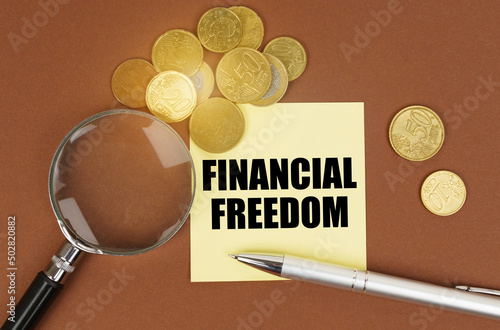On a brown surface are coins, a pen, a magnifying glass and stickers with the inscription - FINANCIAL FREEDOM