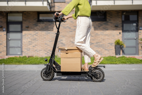 Young happy woman driving electric scooter, carrying cardboard boxes on street in residential area. Concept of sustainability, delivery and eco-friendly modern lifestyle