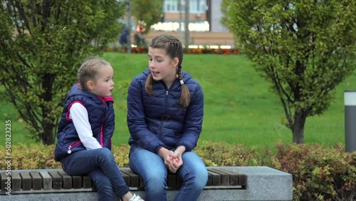 Caucasian girls communicate with each other while sitting on a bench in a public park. The children take their mother by the hand and leave with her. Medium full shot of two sisters during the day. 