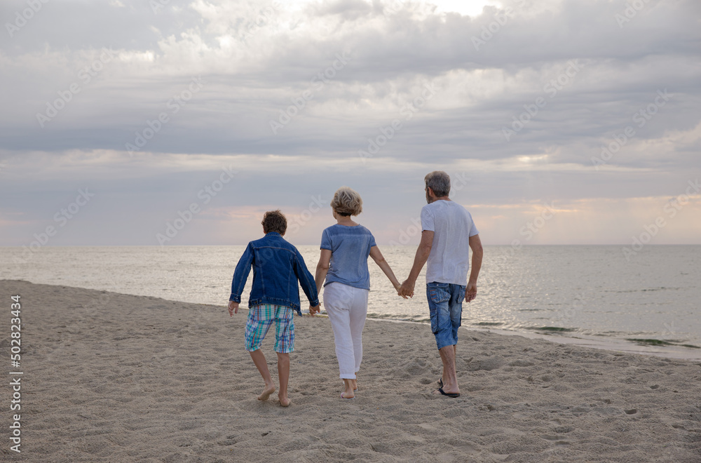 The boy and grandparents hold hands walking by the sea at dawn. Two generations walk by the sea. Morning walk on the beach. Horizontal family view from the back The boy and grandparents hold hands walking by the sea at dawn. Two generations walk by the sea. Morning walk on the beach. Horizontal family view from the back