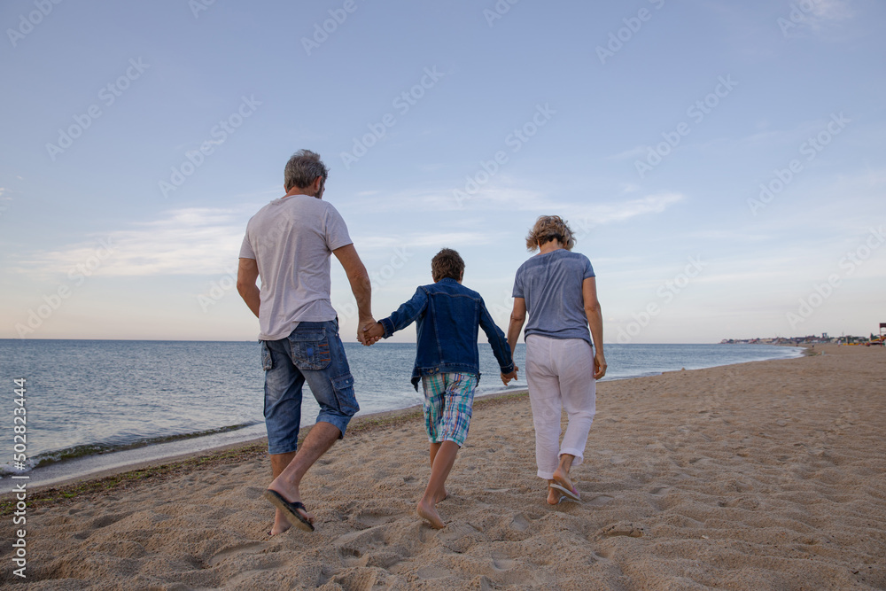 The boy and grandparents hold hands walking by the sea at dawn. Two generations walk by the sea. Morning walk on the beach. Horizontal family view from the back The boy and grandparents hold hands walking by the sea at dawn. Two generations walk by the sea. Morning walk on the beach. Horizontal family view from the back