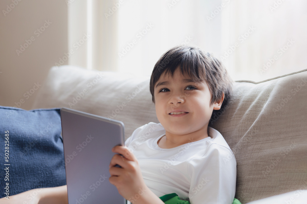 Cinematic portrait young boy playing game on tablet sitting on sofa with light shining from window, Kid playing games online on internet at home, Child talking video call with friends at home Cinematic portrait young boy playing game on tablet sitting on sofa with light shining from window, Kid playing games online on internet at home, Child talking video call with friends at home