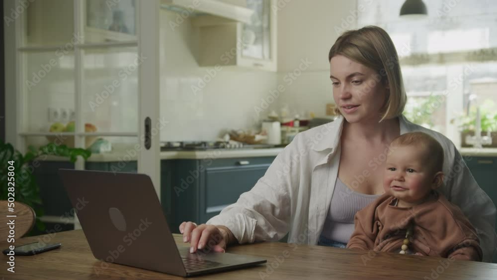 Young adult caucasian mother holding her son and working on laptop in joy Young adult caucasian mother holding her son and working on laptop in joy