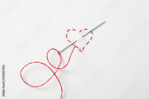 Needle with red thread and stitches in shape of heart on white fabric, top view