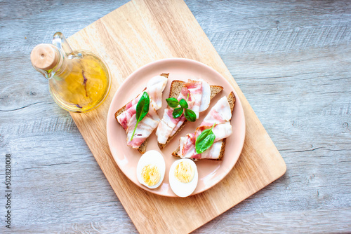 Food, breakfast. Plate with sandwiches and eggs. Rye bread with bacon slice and basil leaf. Olive aromatic oil with pepper. Top view. Flat lay
