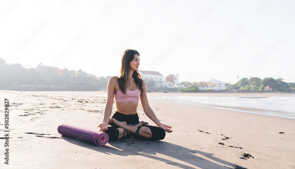 Fototapeta premium Contemplative female sitting in lotus pose and thinking about hatha holistic healing during morning time at coastline beach, pondering Caucasian woman 20s meditate for recreating at seashore