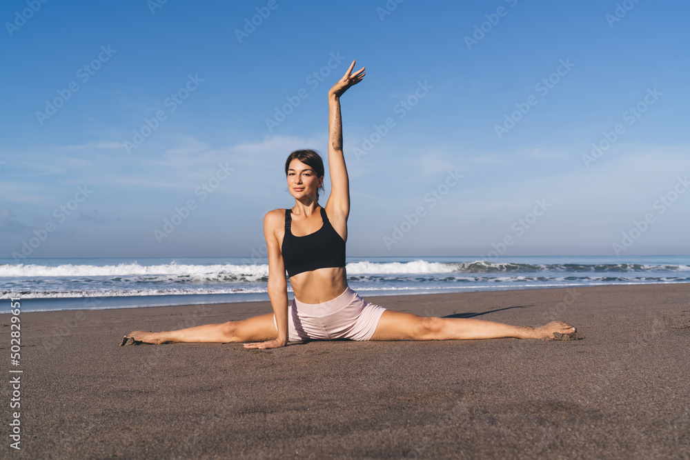 Portrait of young woman sitting in twine doing stretching exercises during workout at seashore beach, experienced female yogi practicing poses for training body flexibility and concentration Portrait of young woman sitting in twine doing stretching exercises during workout at seashore beach, experienced female yogi practicing poses for training body flexibility and concentration