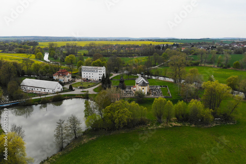 View from above. We see a church with a cemetery  a pond and other buildings. There is a lot of greenery everywhere.