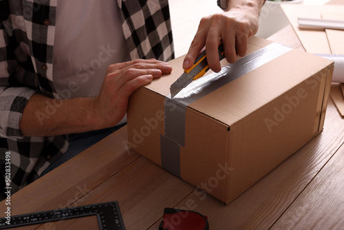 Man using utility knife to open parcel at wooden table, closeup © New Africa