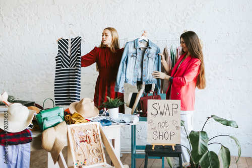 two young woman female caucasian students at swap party try on clothes, bags, shoes and accessories, change clothes with each other, second hand for things, zero waste life, eco-friendly photo