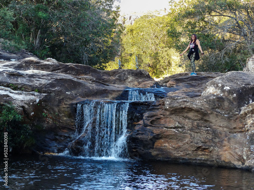 Pequena Cachoeira entre pedras, com muita vegetação em volta e mulher passeiando, localizada na região rural de Três Barras, município do Serro, Minas Gerais, Brasil.