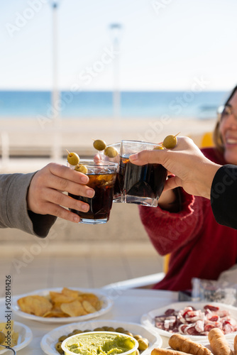 Thre people toasting with vermouth in front of the beach in a terrace. One young woman sharing snacks with friends outdoor during a sunny day.