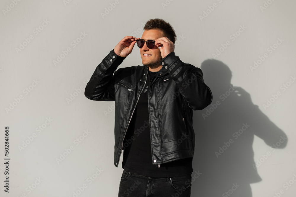 Cool brutal hipster man with a smile wearing fashionable black clothes with a leather jacket wears black glasses on a gray background in the city Cool brutal hipster man with a smile wearing fashionable black clothes with a leather jacket wears black glasses on a gray background in the city