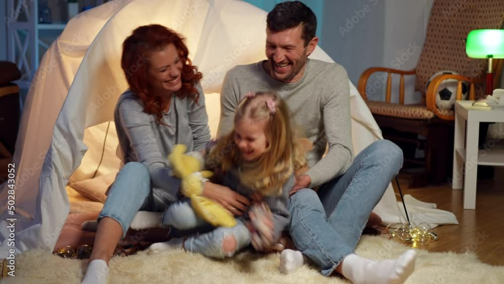 Joyful daughter running to parents sitting at tent in living room at home indoors. Happy cheerful little Caucasian girl playing with father and mother having fun. Laughing man and woman tickling child Joyful daughter running to parents sitting at tent in living room at home indoors. Happy cheerful little Caucasian girl playing with father and mother having fun. Laughing man and woman tickling child
