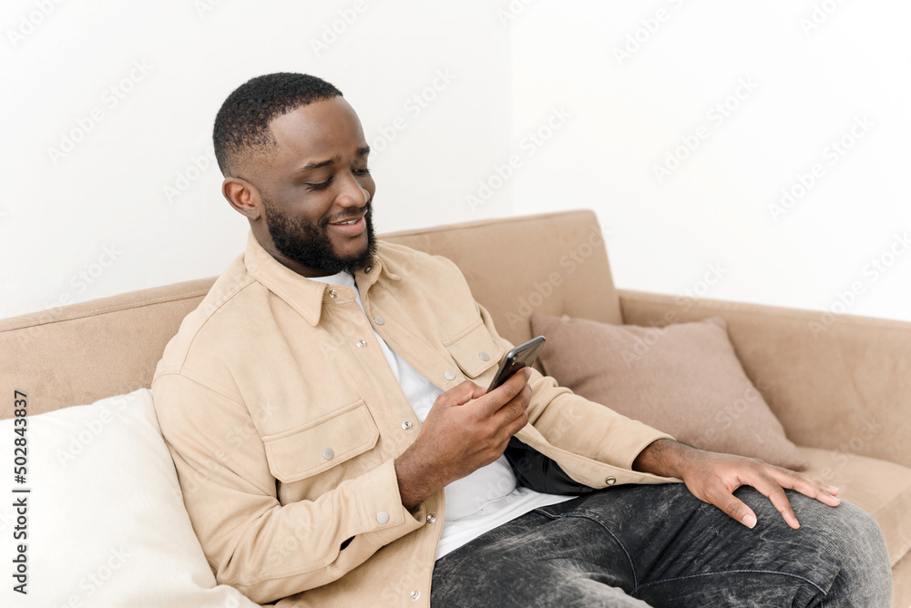 Smiling african american man browsing news or social media feed on phone while sitting on sofa at home. Rest and relaxation