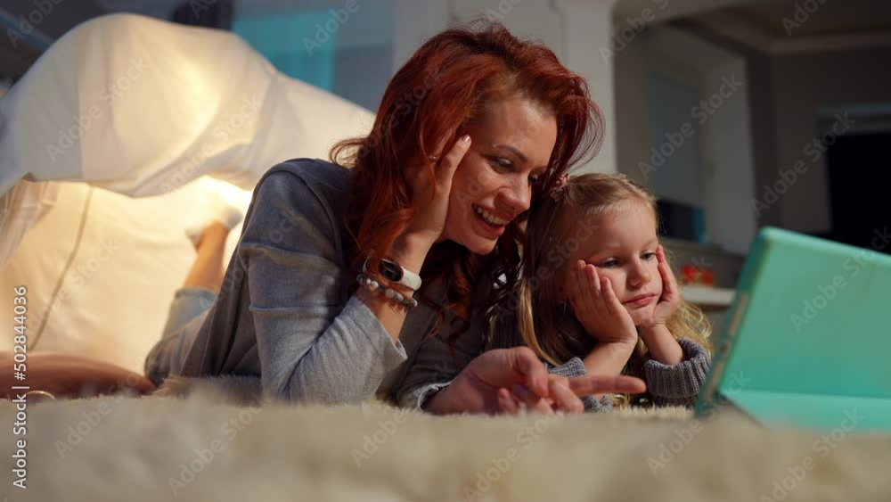 Positive Caucasian mother and daughter talking watching cartoon online on tablet. Relaxed woman and girl lying at tent in living room discussing film smiling. Family leisure and happiness concept Positive Caucasian mother and daughter talking watching cartoon online on tablet. Relaxed woman and girl lying at tent in living room discussing film smiling. Family leisure and happiness concept