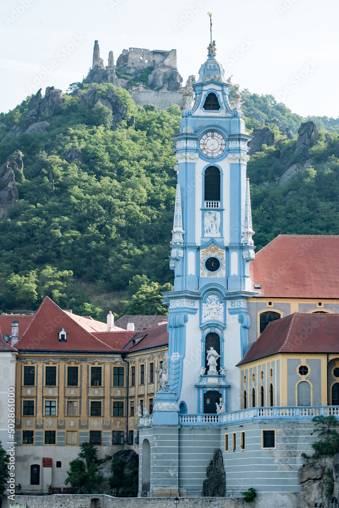 The blue and white tower of Stift Durnstein rises next to the Danube River and a 12th century medieval castle, Burgruine Dürnstein, looms behind on a mountainside.