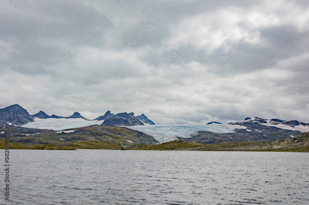 Norwegian mountain landscape with snow covered melting  glacier, lake and grey sky with clouds. 