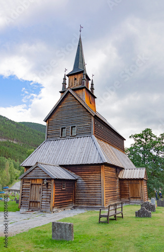 Kaupanger Stave Church in western Norway. Old wooden walls and bell tower, cloudy grey sky, green grass and trees, stone tombs of the old cemetery, mountain slopes on the background