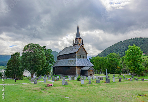 Kaupanger Stave Church in western Norway. Old wooden walls and bell tower, cloudy grey sky, green grass and trees, stone tombs of the old cemetery, mountain slopes on the background