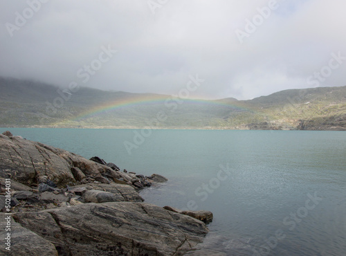 Rainbow in the cloud above the  Styggevatnet glacier lake in Norway. Rocky shore and foggy mountains around