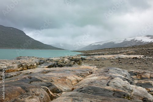 Shore of the Styggevatnet glacier lake in Norway. Turquoise water, grey and brown stones, snor on the mountain slopes, cloudy sky